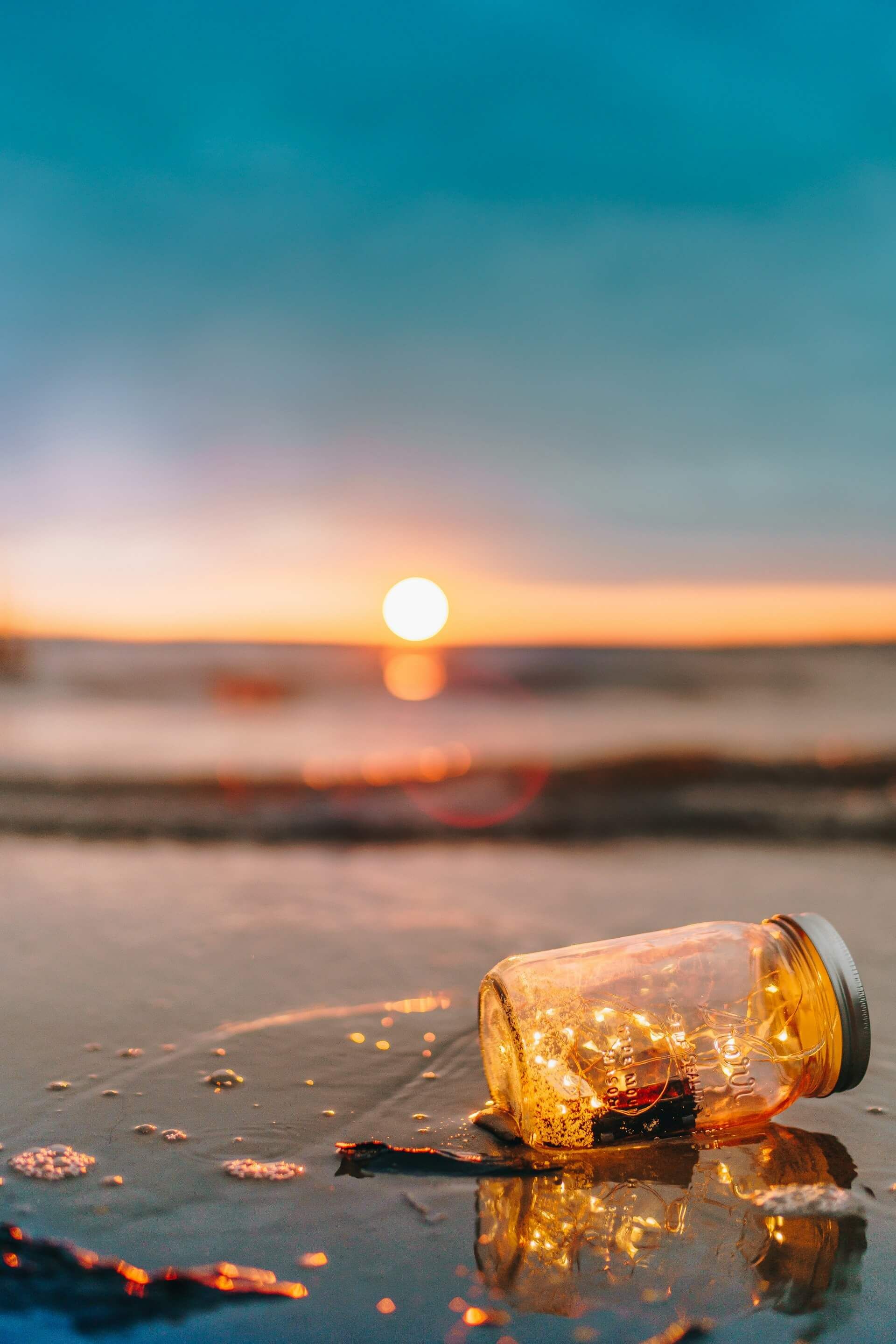 Bottle with fairy lights on a wet beach, illuminated by a vivid ocean sunset.