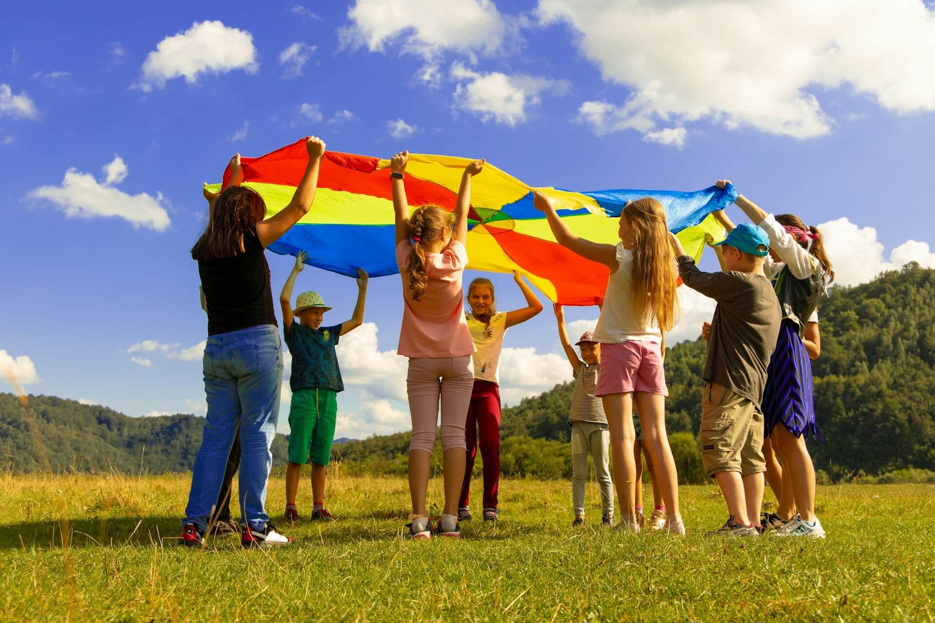 Group of children holding a colorful parachute up in a sunny, grassy field.