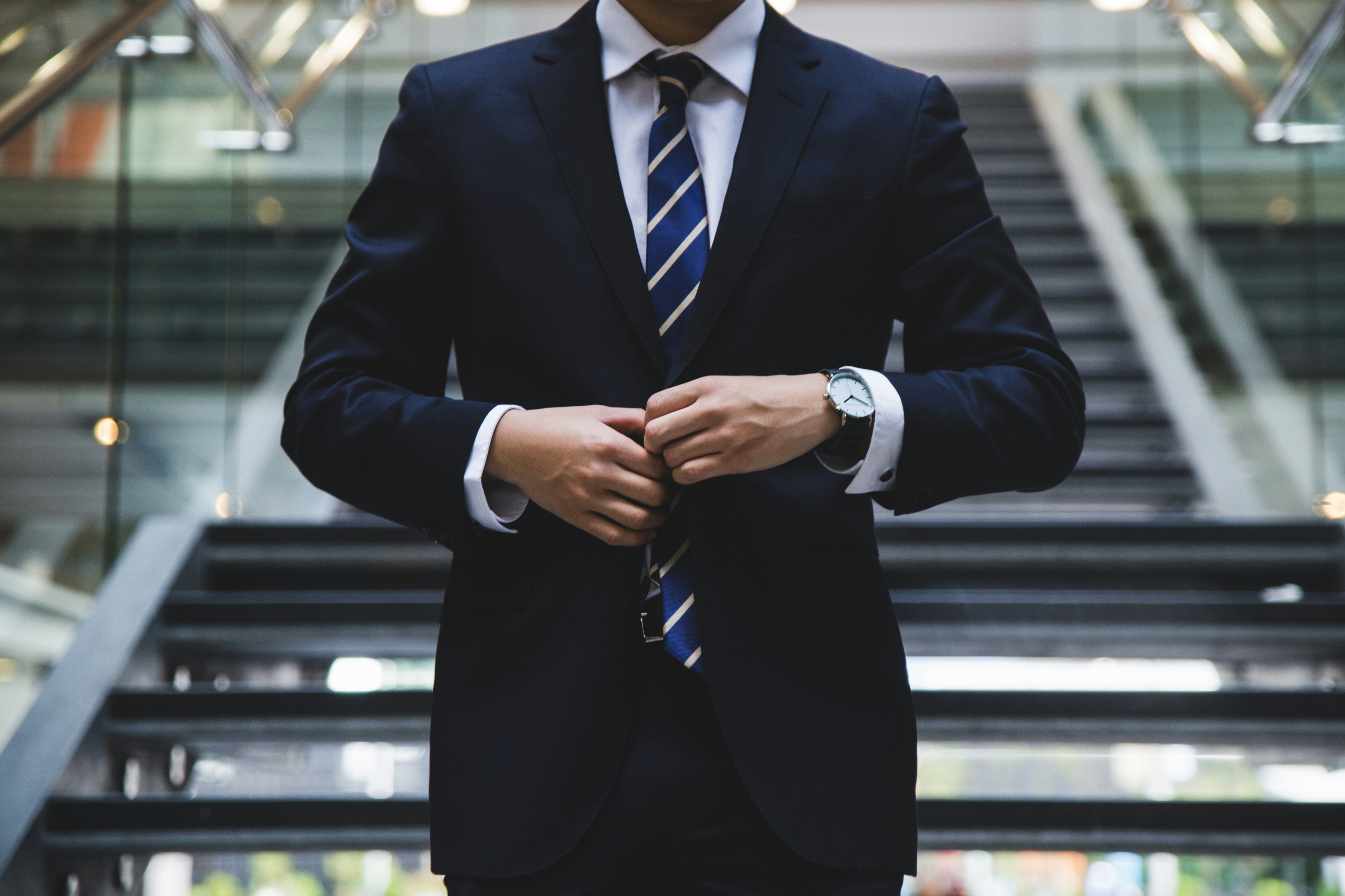 Well-dressed man in a dark suit, white shirt, and striped tie adjusting his jacket lapel indoors.
