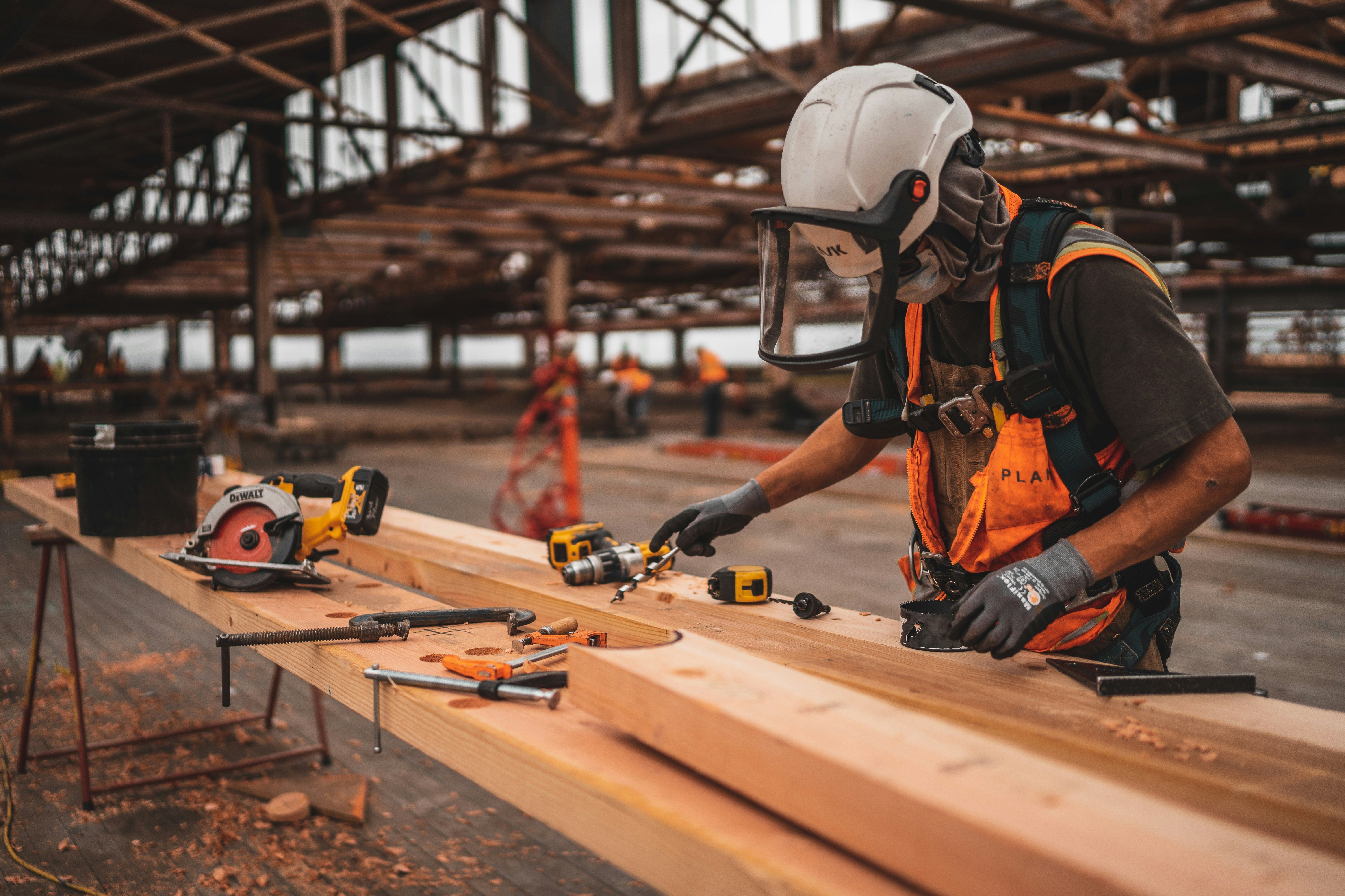 Construction worker in safety gear shaping wooden beams with various tools on a workbench.