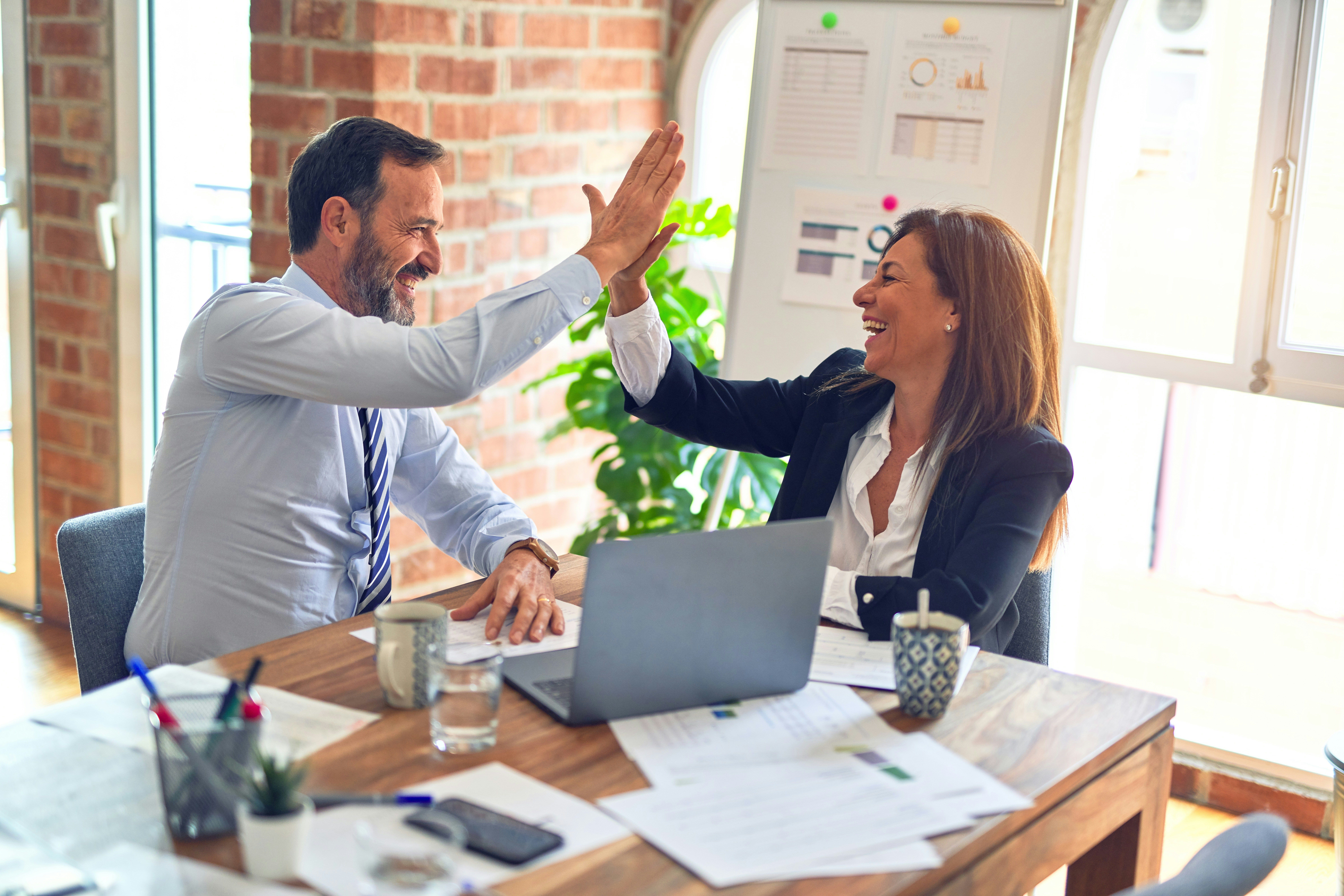 Two smiling colleagues high-five across a desk in a bright office, celebrating success.