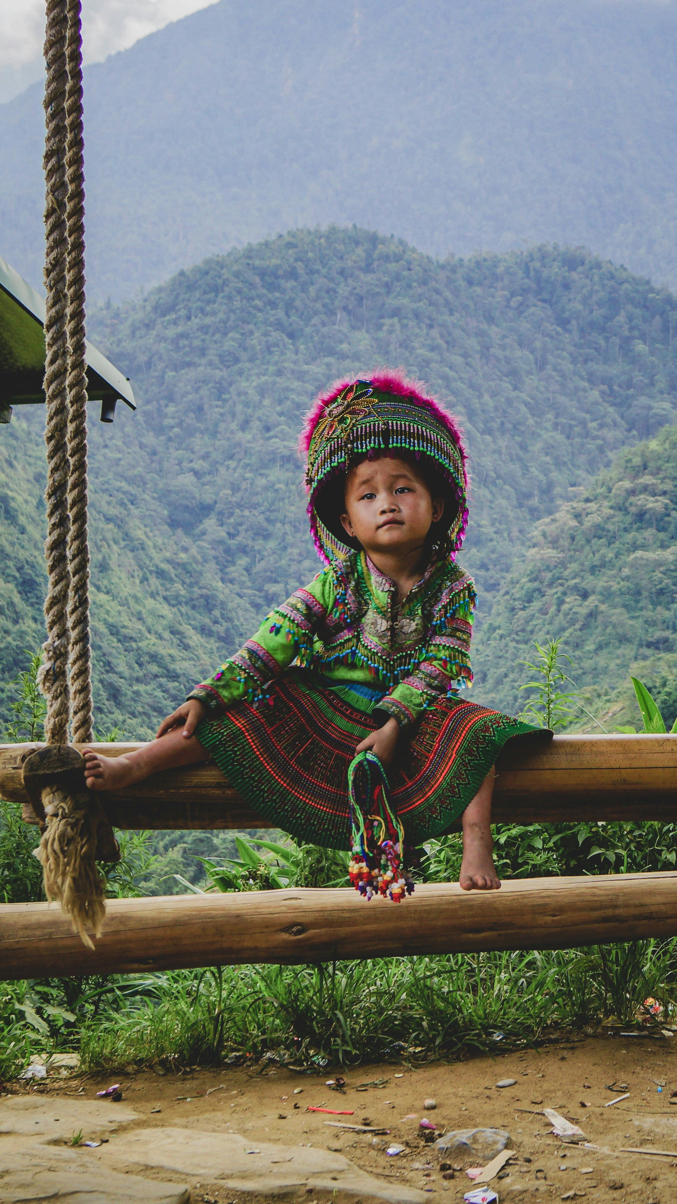 A young child in traditional embroidered clothing sits on a wooden rail with lush green mountains behind.
