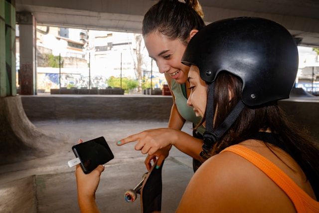 Two women at a skatepark looking at a smartphone, one wearing a black helmet.